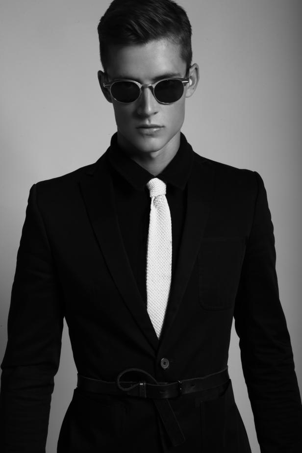 Ein Mann mit Sonnenbrille, schwarzem Anzug und weißer Strickkrawatte.