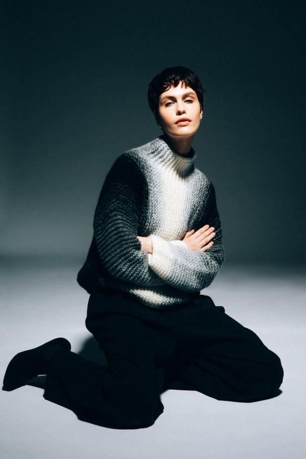 Eine Frau kniet mit verschränkten Armen und trägt einen grob gestrickten Pullover.