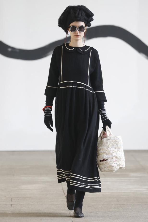 Eine Frau mit Hut und Sonnenbrille trägt ein schwarzes Kleid mit weißen Akzenten und eine Tasche.