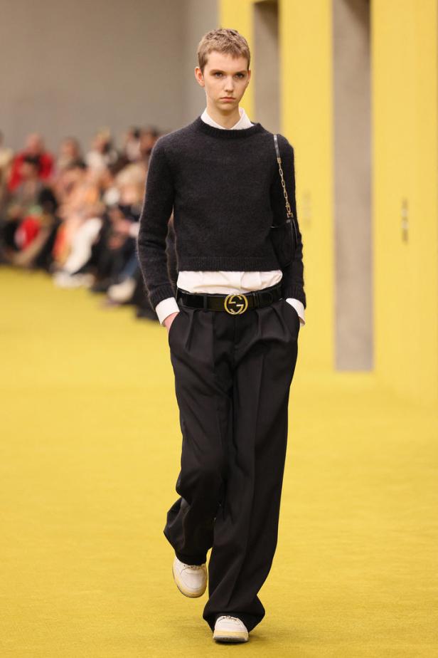 Ein Model läuft in einem schwarzen Pullover und einer Hose mit Gucci-Gürtel über den Laufsteg.