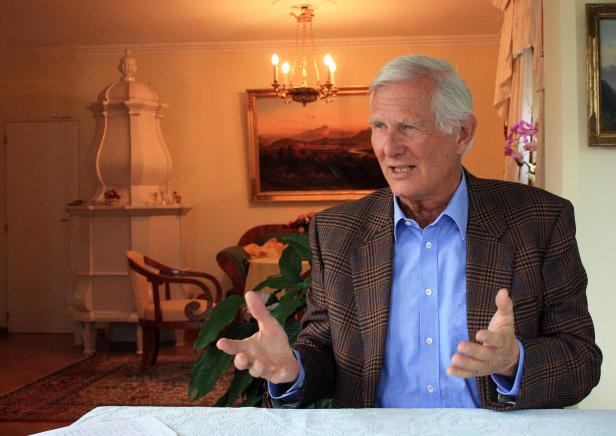 Ein älterer Mann mit blauem Hemd und kariertem Sakko gestikuliert in einem Wohnzimmer.