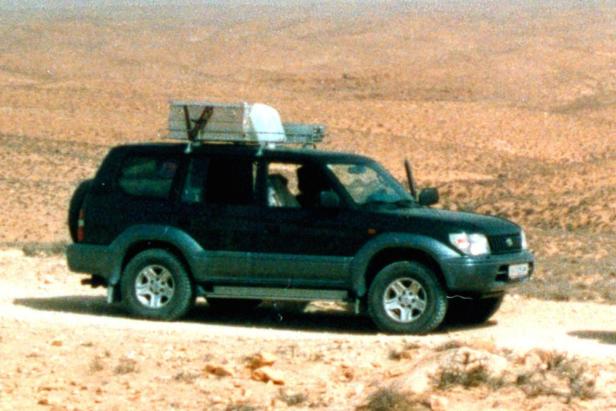 Ein dunkler Geländewagen mit Gepäck auf dem Dach fährt durch eine Wüstenlandschaft.