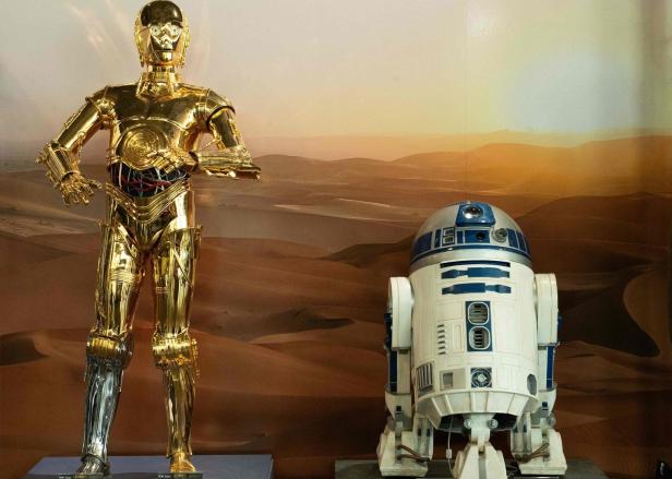 Die Droiden C-3PO und R2-D2 aus „Star Wars“ stehen vor einer Wüstenlandschaft.