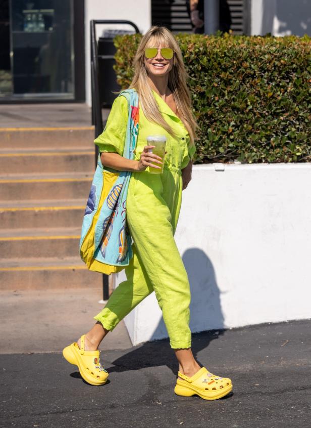 Heidi Klum in einem hellgrünen Overall, gelben Crocs und mit einer bunten Tasche über der Schulter.