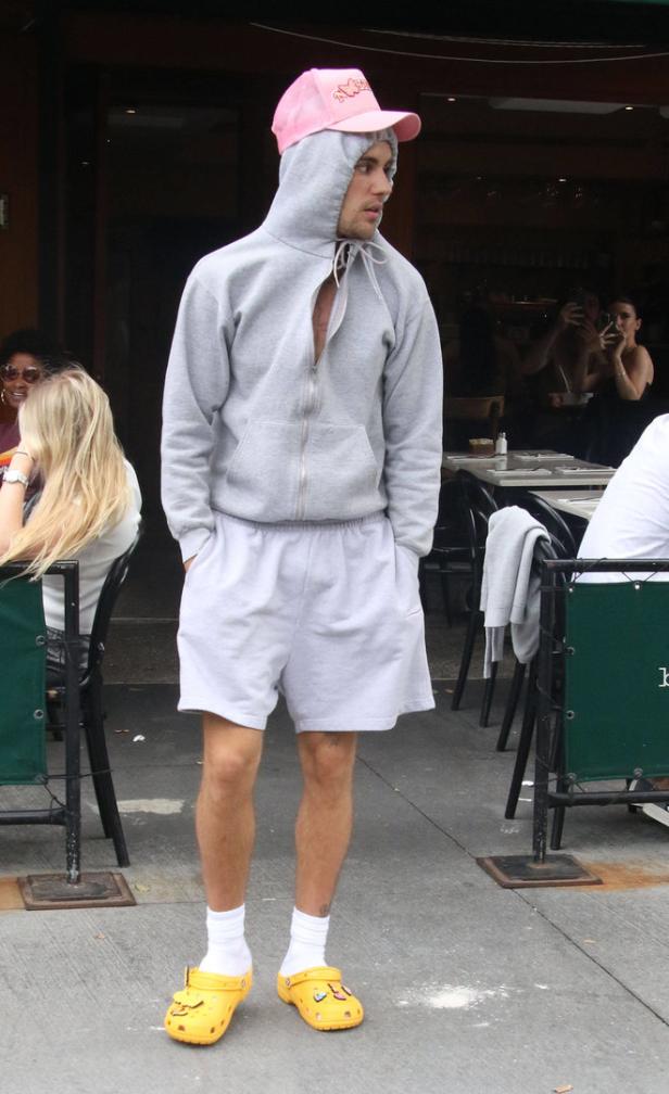 Ein Mann mit rosa Kappe, Shorts und gelben Crocs steht vor einem Café.