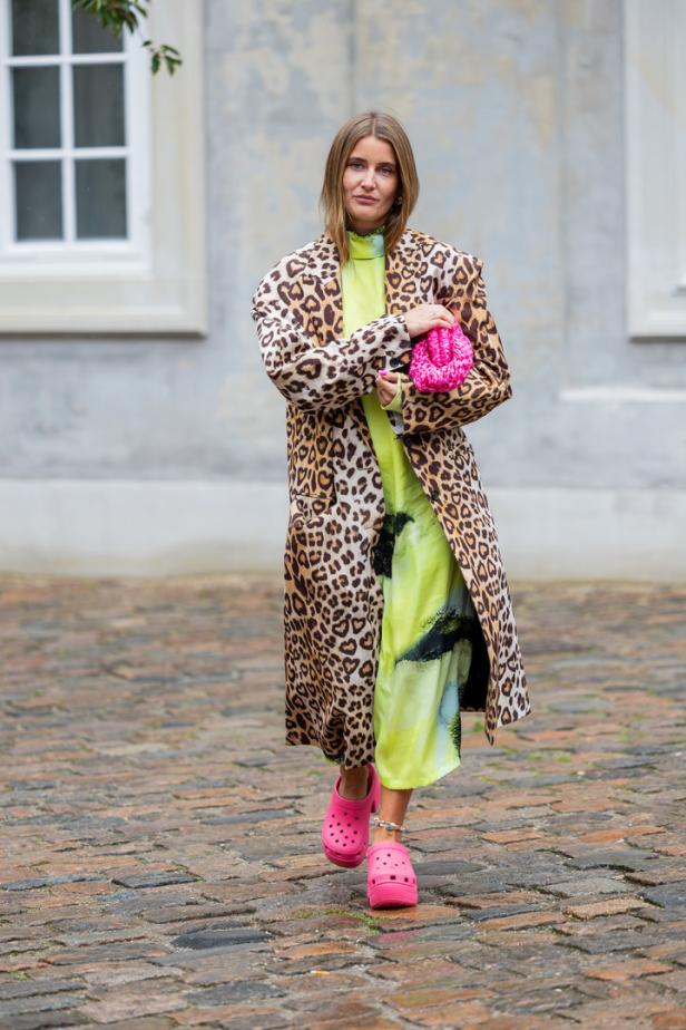 Eine Frau mit Leopardenmantel, neonfarbenem Kleid und pinkfarbenen Crocs posiert auf einem gepflasterten Weg.