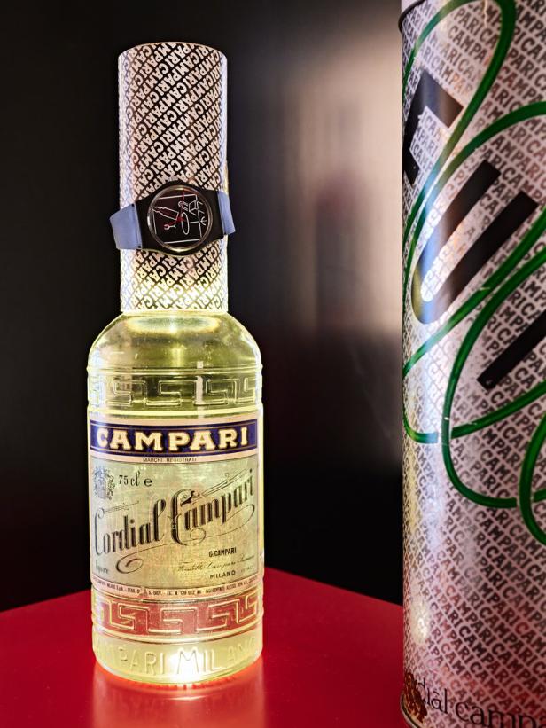 Eine Flasche Cordial Campari mit einer Swatch-Uhr am Hals auf einem roten Tisch.