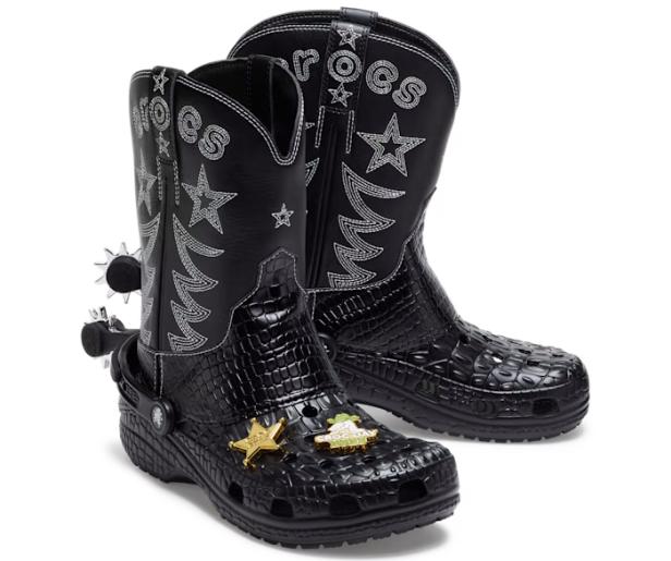 Zwei schwarze Crocs-Cowboystiefel mit Sporen und goldenen Ansteckern stehen auf einem weißen Hintergrund.