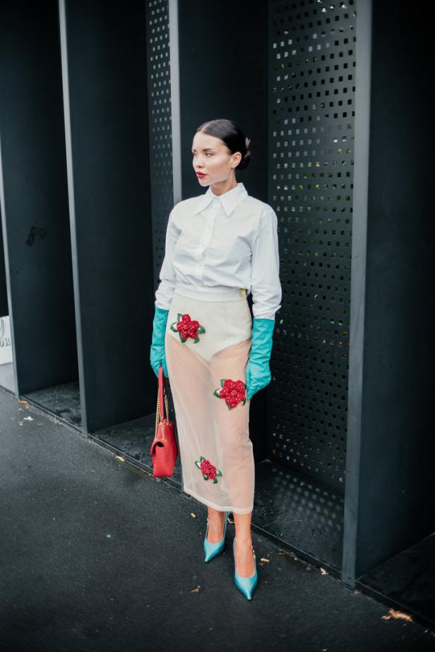 Eine Frau in einem weißen Hemd, einem transparenten Rock mit Blumenapplikationen und blauen Handschuhen und Schuhen.