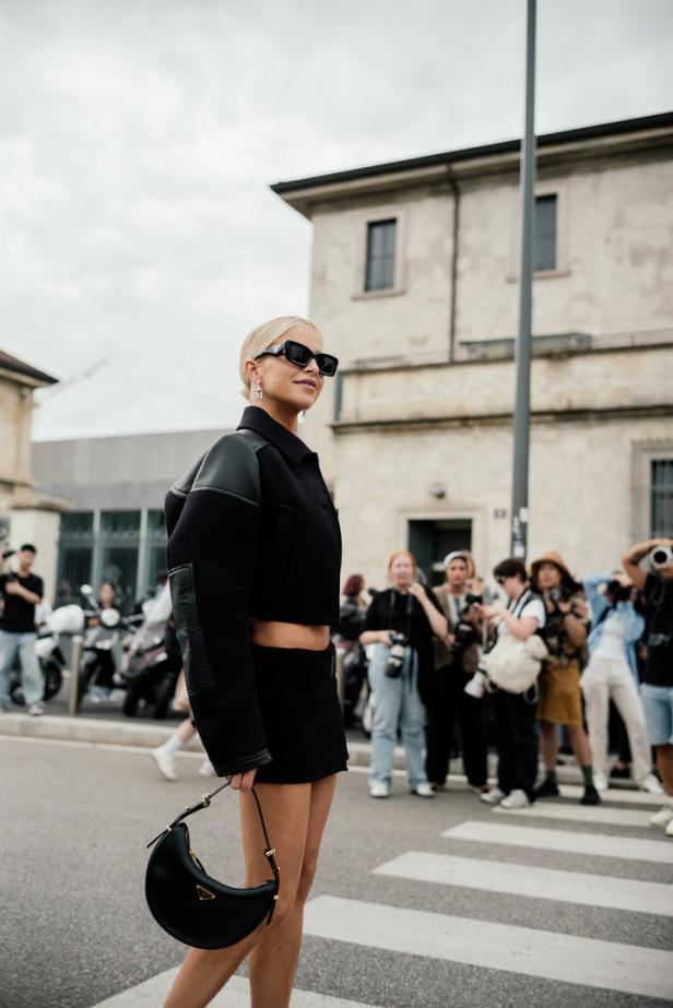 Eine Frau mit Sonnenbrille und schwarzem Outfit überquert eine Straße.