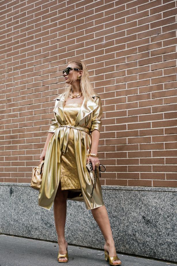Eine Frau in einem goldenen Outfit posiert vor einer Backsteinmauer.