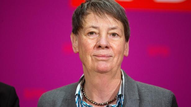 Eine Frau mit kurzem braunem Haar und einer Perlenkette vor einem pinkfarbenen Hintergrund.