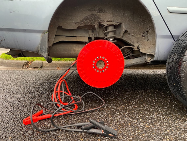Ein Auto mit abmontiertem Rad und einem roten Bremsscheibenschutz sowie Starthilfekabeln.