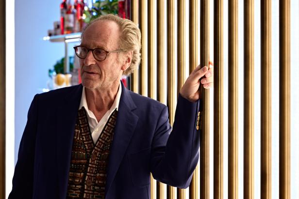 Ein älterer Mann mit Brille, gekleidet in Sakko und Weste, steht vor einer Wand mit goldenen Streifen.