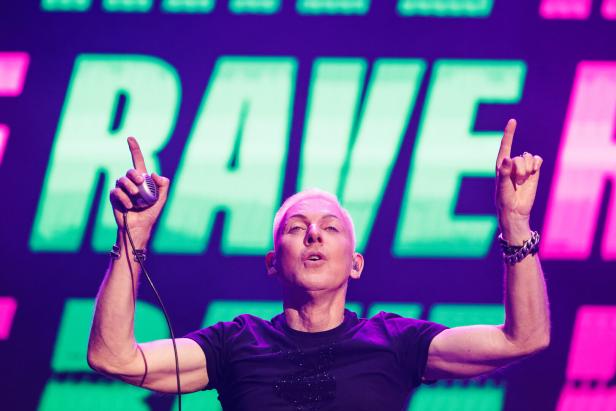 H. P. Baxxter mit erhobenen Händen steht vor einer großen „RAVE“-Leuchtschrift.