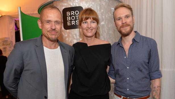Drei Personen posieren vor einer Wand mit dem Schriftzug „Brothers“.