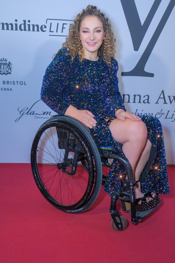 Kristina Vogel mit lockigem Haar sitzt in einem Rollstuhl und trägt ein blaues, glitzerndes Abendkleid vor einer Sponsorenwand.