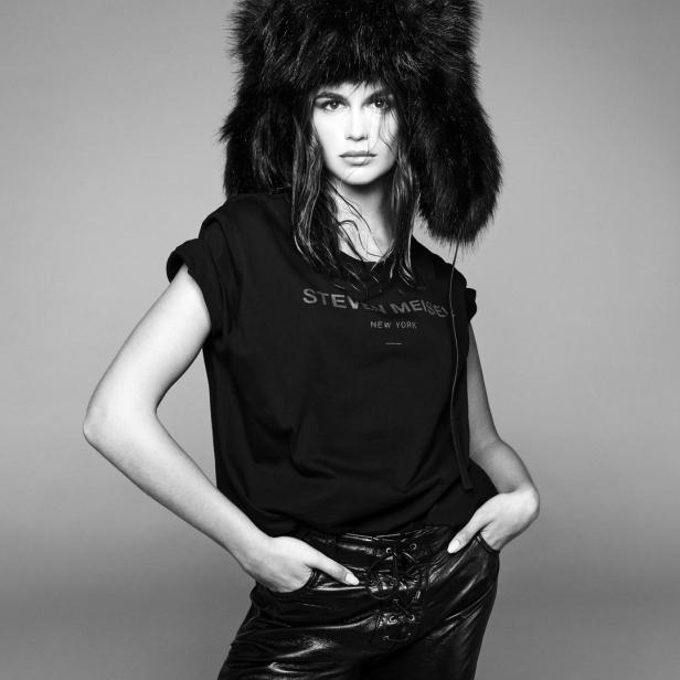 Eine Frau mit Pelzmütze und einem T-Shirt mit der Aufschrift „Steven Meisel New York“ posiert in Lederhosen.