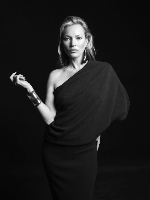 Kate Moss in einem schwarzen, schulterfreien Kleid und auffälligem Schmuck vor schwarzem Hintergrund.