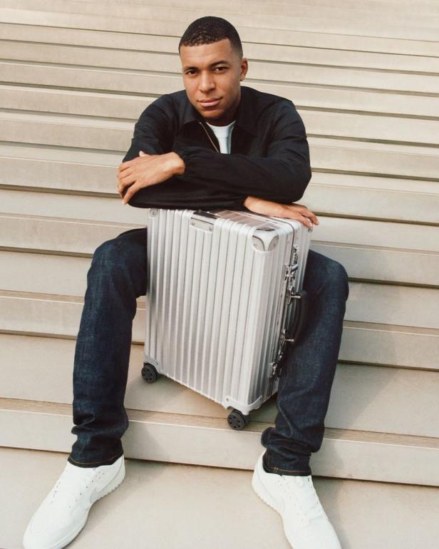 Kylian Mbappé sitzt mit einem silbernen Rollkoffer auf einer Treppe.