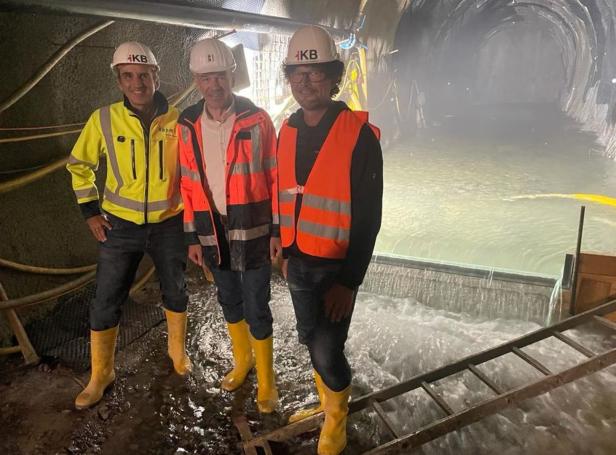 Drei Männer, darunter Bürgermeister Willi, stehen mit Sicherheitsausrüstung vor einem mit Wasser gefüllten Tunnel.