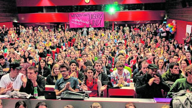 Ein vollbesetzter Hörsaal mit Studierenden und einem Banner mit der Aufschrift „Wessen Uni? Unsere Uni!“.