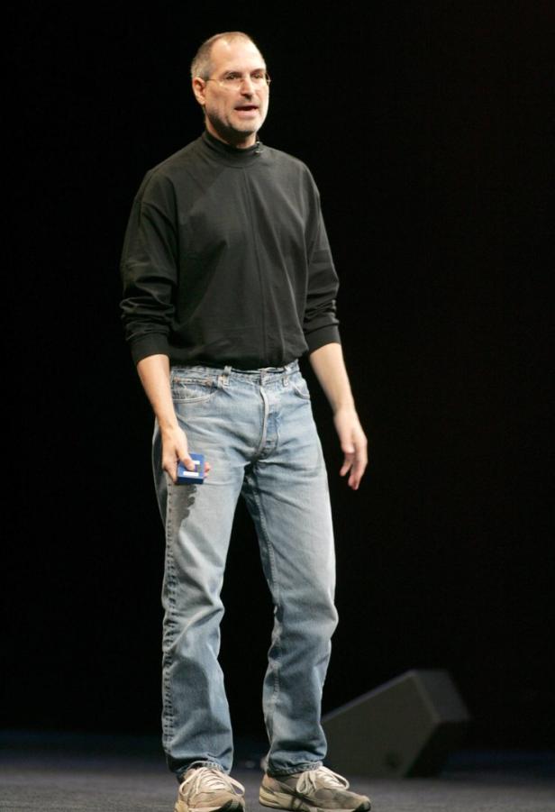 Steve Jobs steht auf einer Bühne und hält ein Gerät in der Hand.