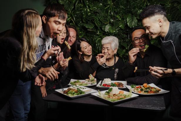 Eine Gruppe von Menschen isst gemeinsam asiatische Speisen an einem Tisch vor einer grünen Wand.