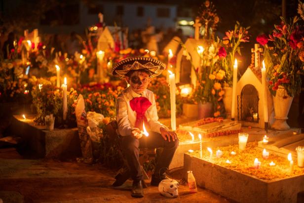 Ein Junge mit Sombrero und Gesichtsbemalung sitzt auf einem Friedhof, der mit Blumen und Kerzen geschmückt ist.