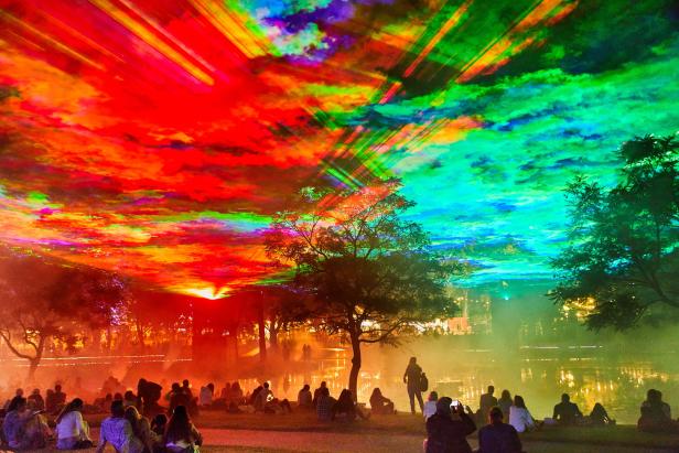 Zuschauer beobachten eine farbenfrohe Lichtshow über einem See.