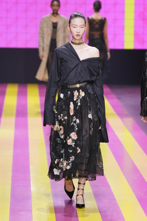 Ein Model präsentiert ein schwarzes Hemd und einen Rock mit Blumenmuster auf dem Laufsteg.