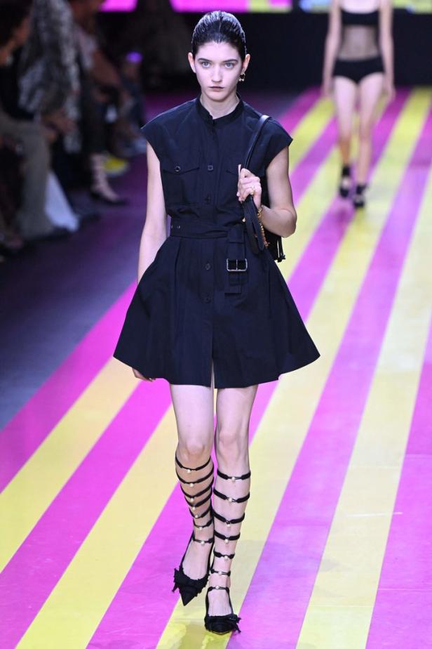 Ein Model präsentiert ein schwarzes Kleid und Gladiatorensandalen auf einem Laufsteg.