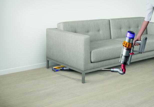 Eine Person saugt mit einem Dyson Staubsauger unter einem grauen Sofa.