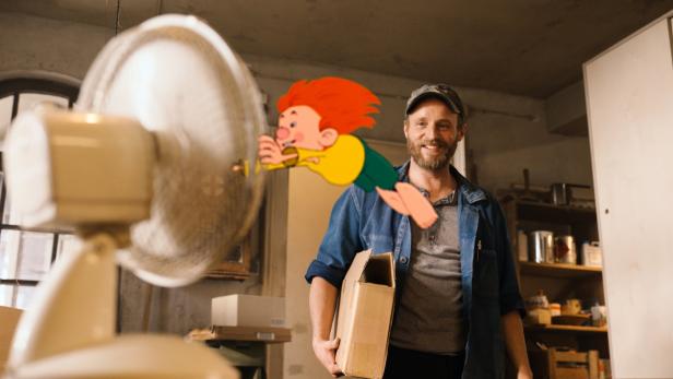 Ein Mann mit Bart hält einen Karton, während ein rothaariger Comic-Junge durch einen Ventilator fliegt.