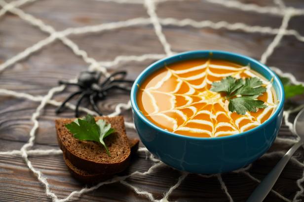 Eine Kürbissuppe mit Spinnennetz-Dekoration in einer blauen Schüssel, serviert mit Brot.
