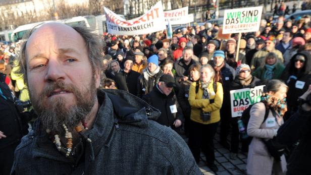 Ein Mann mit Bart steht vor einer Demonstration mit Schildern und Bannern.