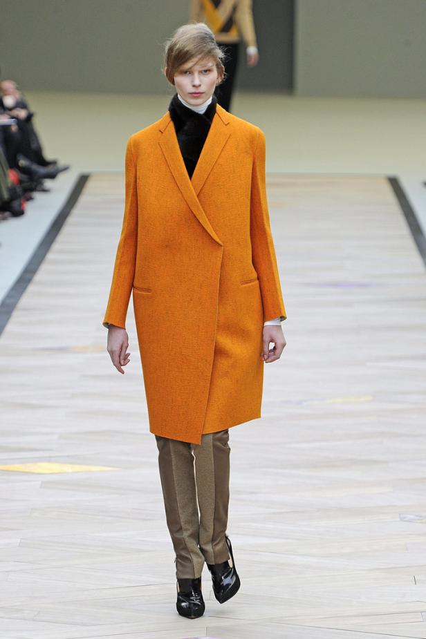 Eine Frau in einem orangefarbenen Mantel und braunen Hosen auf einem Laufsteg.