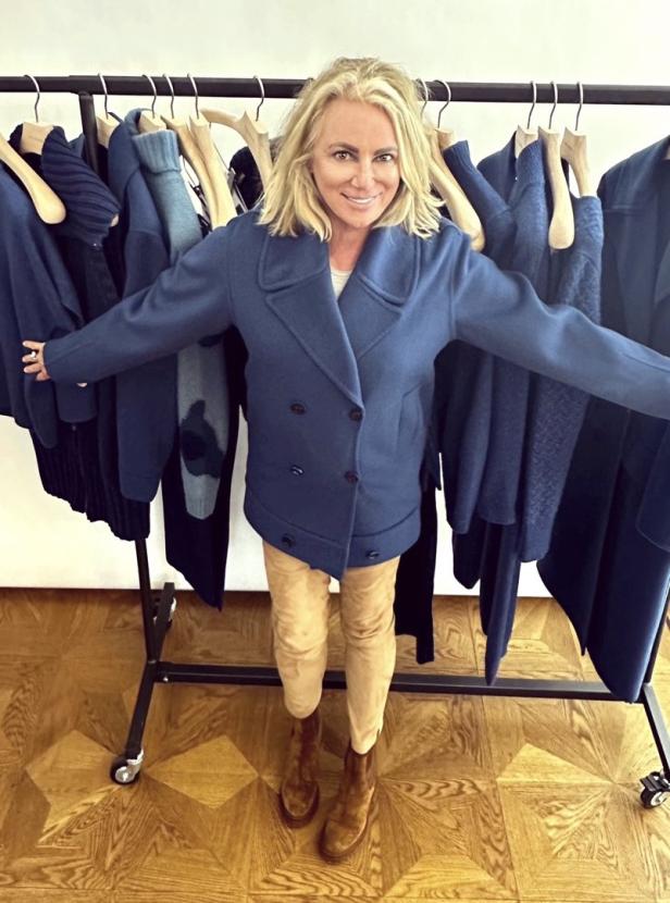 Eine Frau mit blonder Mähne posiert vor einer Kleiderstange mit blauer Kleidung.