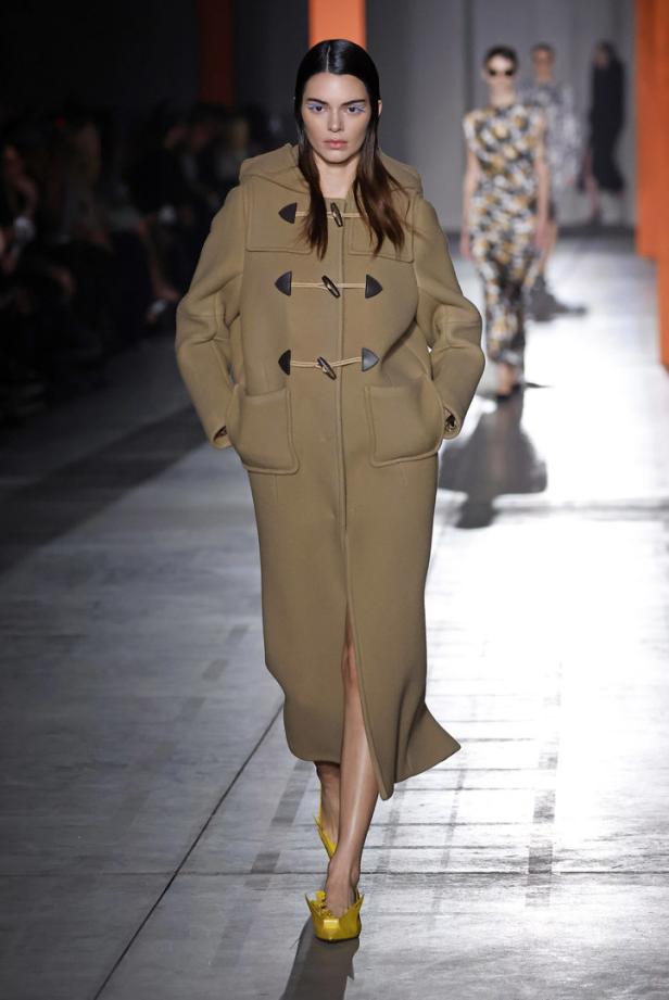 Kendall Jenner läuft in einem langen, beigen Dufflecoat und gelben Schuhen über einen Laufsteg.