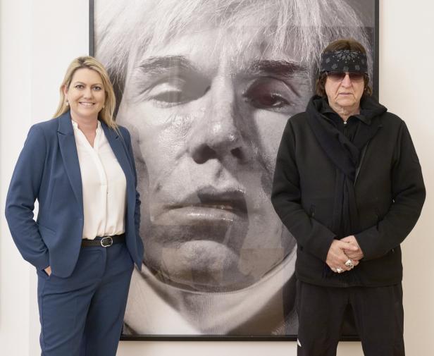 Eine Frau im Hosenanzug und ein Mann mit Stirnband stehen vor einem Porträt von Andy Warhol.