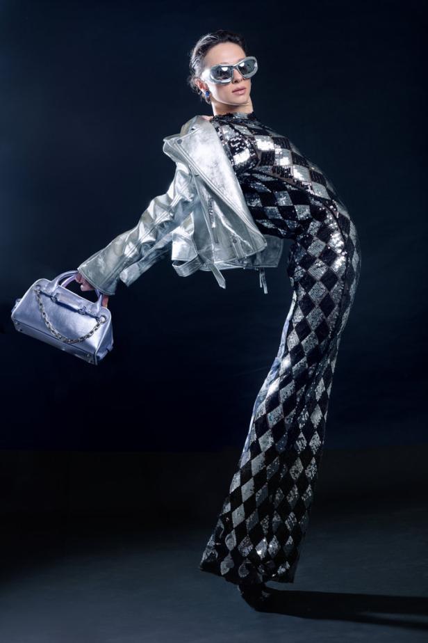 Eine Frau in einem Paillettenkleid mit Rautenmuster, einer silbernen Jacke und futuristischer Sonnenbrille posiert mit einer Handtasche.