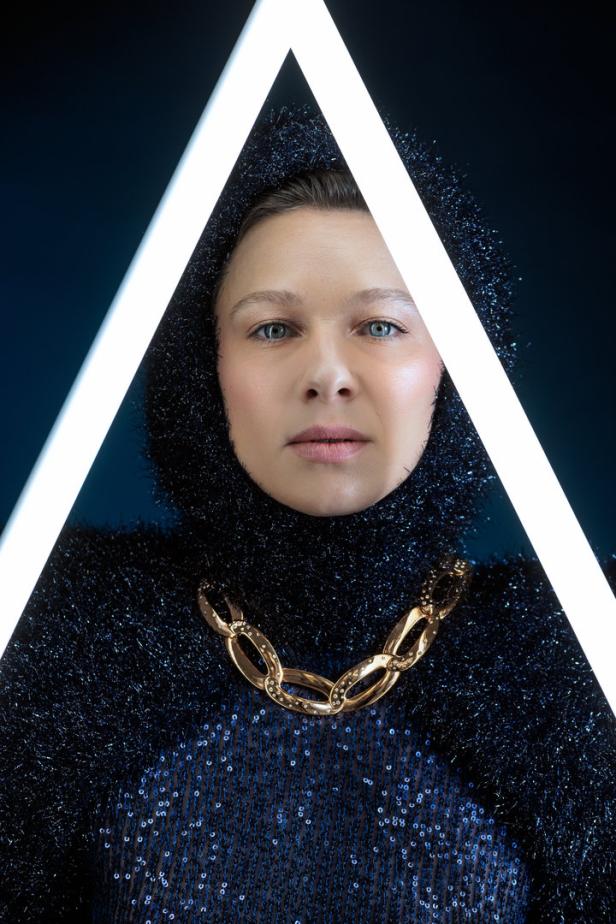 Eine Frau mit Kapuze und goldener Kette steht vor einem dreieckigen Licht.