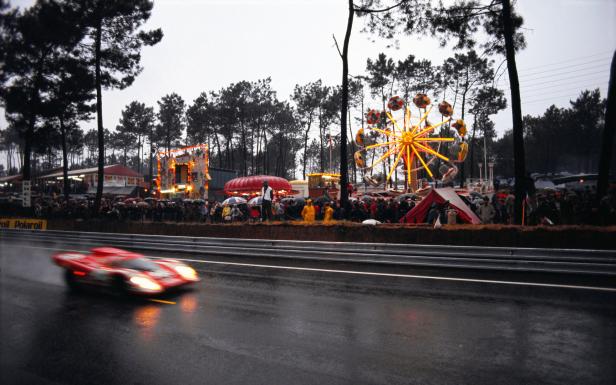 Ein roter Rennwagen fährt an einem Jahrmarkt mit Riesenrad vorbei.
