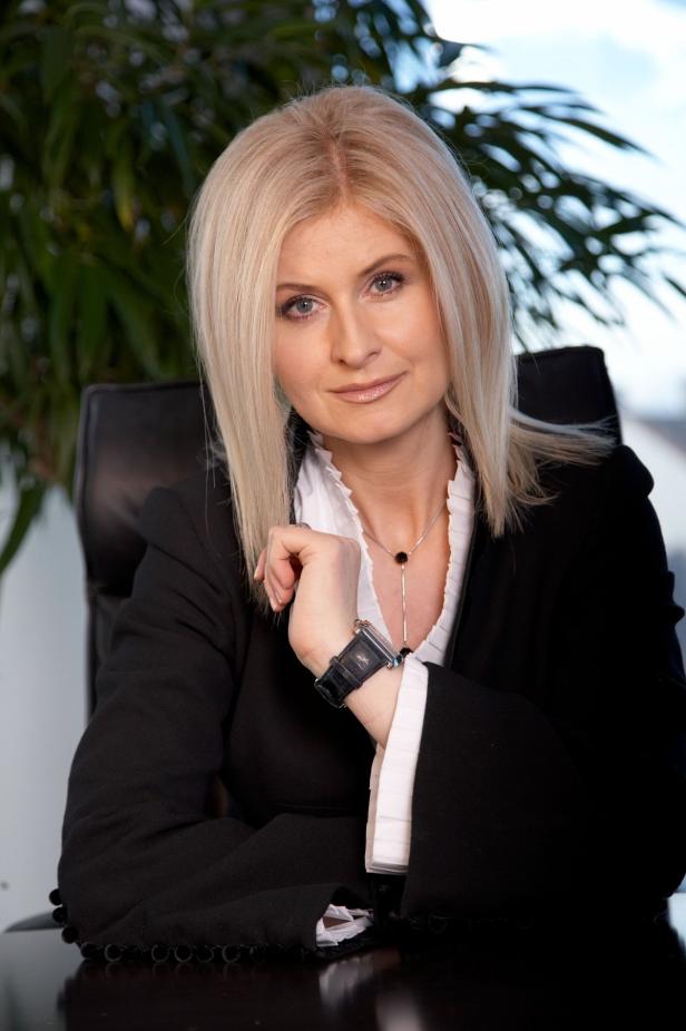 Eine blonde Frau in einem schwarzen Blazer sitzt an einem Schreibtisch.