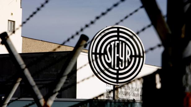 Das Steyr-Firmenlogo vor einem Gebäude, teilweise durch einen Zaun verdeckt.