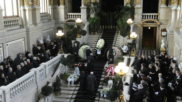 Eine Trauerfeier mit vielen Menschen und Blumengebinden auf einer Treppe.