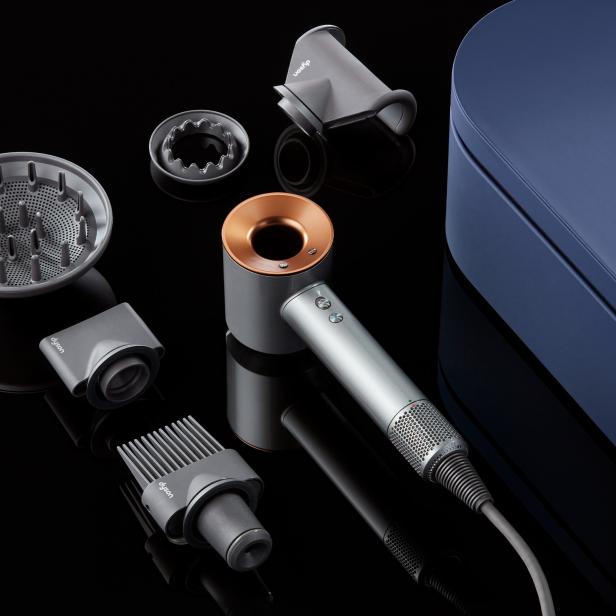 Ein Dyson-Haartrockner mit verschiedenen Aufsätzen und einer blauen Aufbewahrungsbox auf schwarzem Hintergrund.