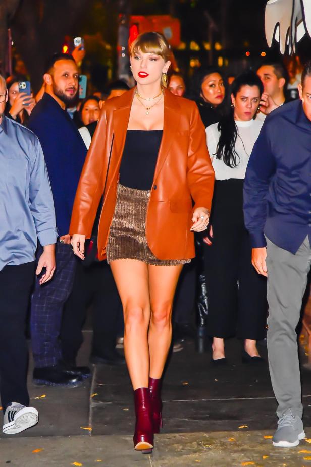 Taylor Swift trägt eine braune Lederjacke, Shorts und rote Stiefel.