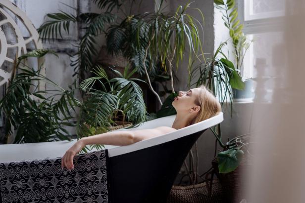 Eine blonde Frau entspannt in einer Badewanne, umgeben von Zimmerpflanzen.
