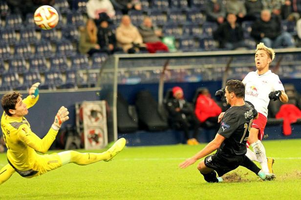 Ein Fußballtorwart in gelber Kleidung springt, um einen Ball abzuwehren, während ein anderer Spieler zusieht.